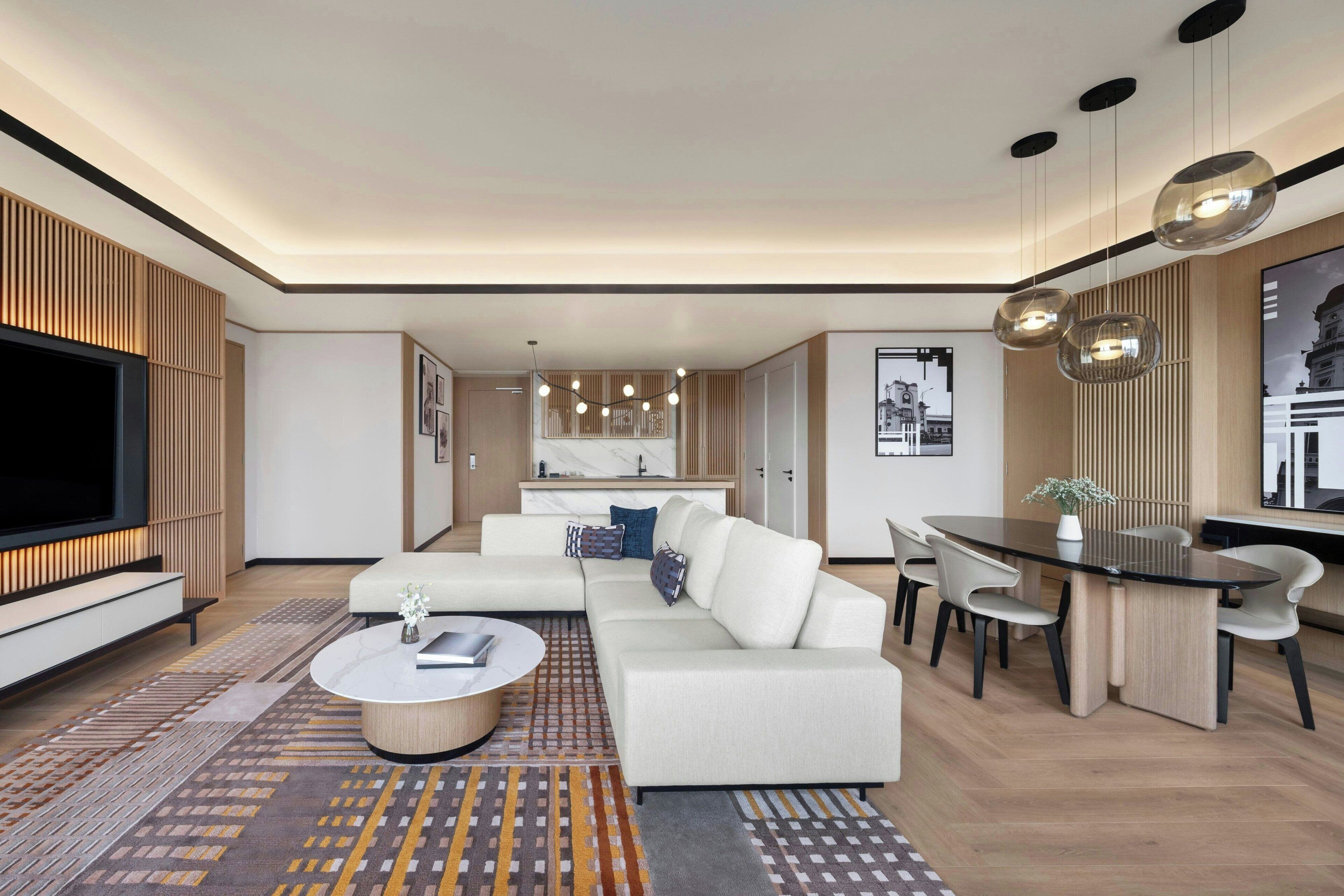 A modern living room with a sectional sofa, a coffee table, and a TV. There is a dining area with a table and chairs. The room has a contemporary design with wooden accents and a patterned rug.