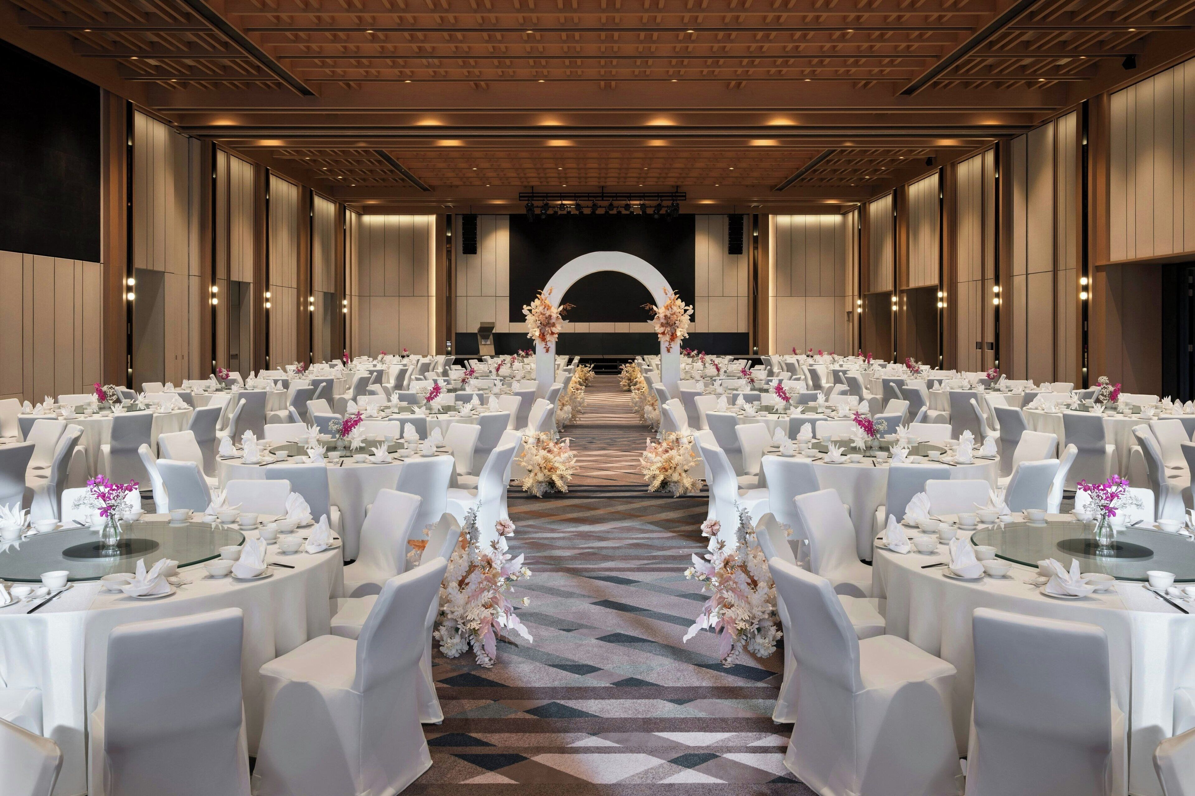 Uma sala de banquetes com mesas redondas, cadeiras brancas e centros de mesa florais.