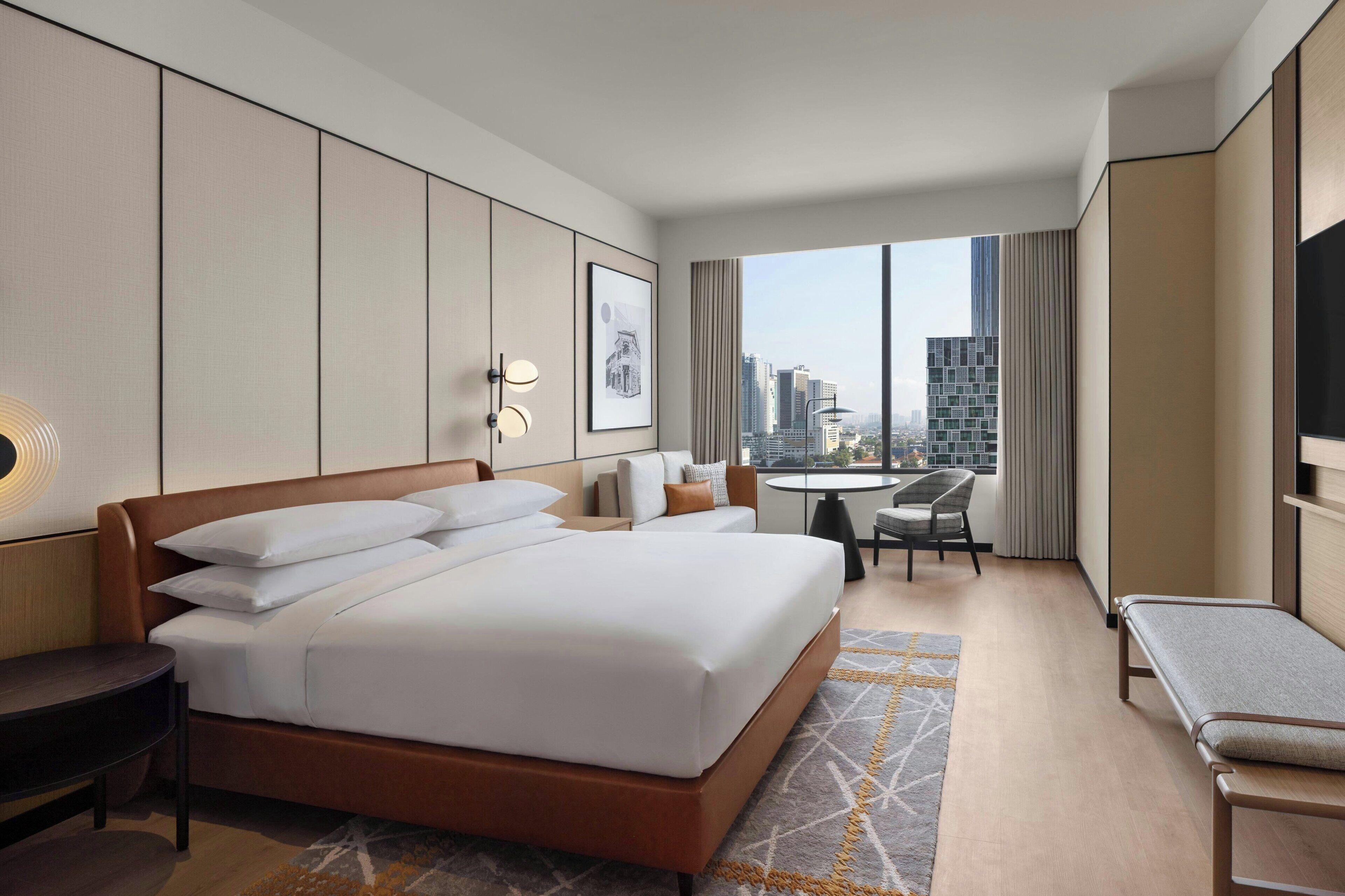 Ein Hotelzimmer mit einem großen Bett, einem Schreibtisch, einem Stuhl und einem kleinen Tisch. Durch das Fenster ist die Stadt zu sehen.