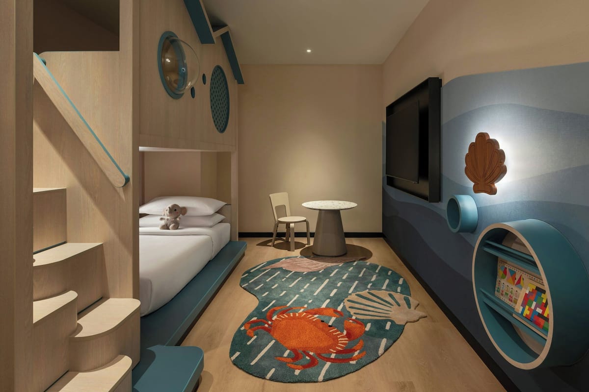 Une chambre d’hôtel compacte comprenant un lit, une petite table, une chaise, un téléviseur et une bibliothèque circulaire originale.