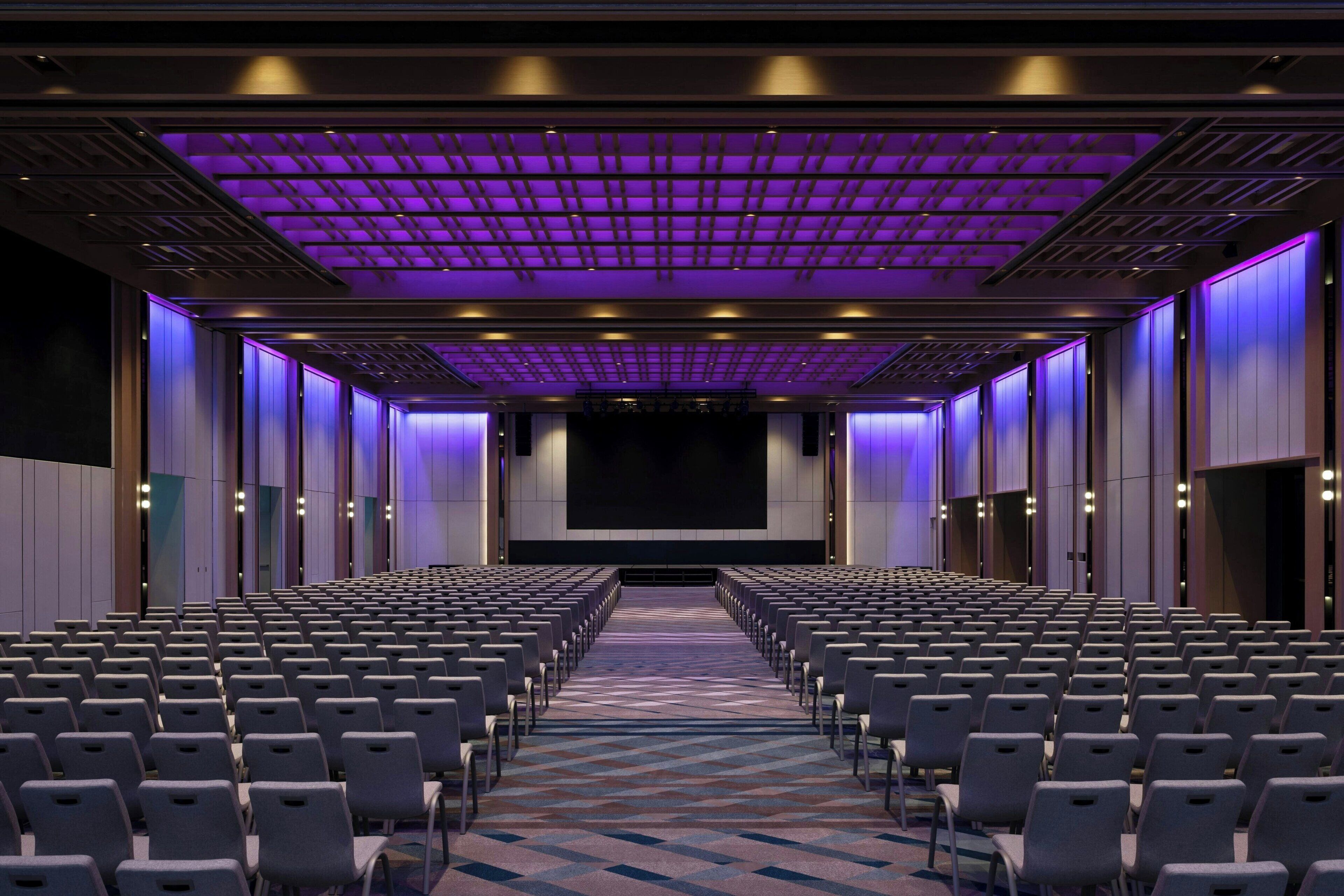 A conference room with rows of chairs, a large screen at the front, and purple lighting on the ceiling.