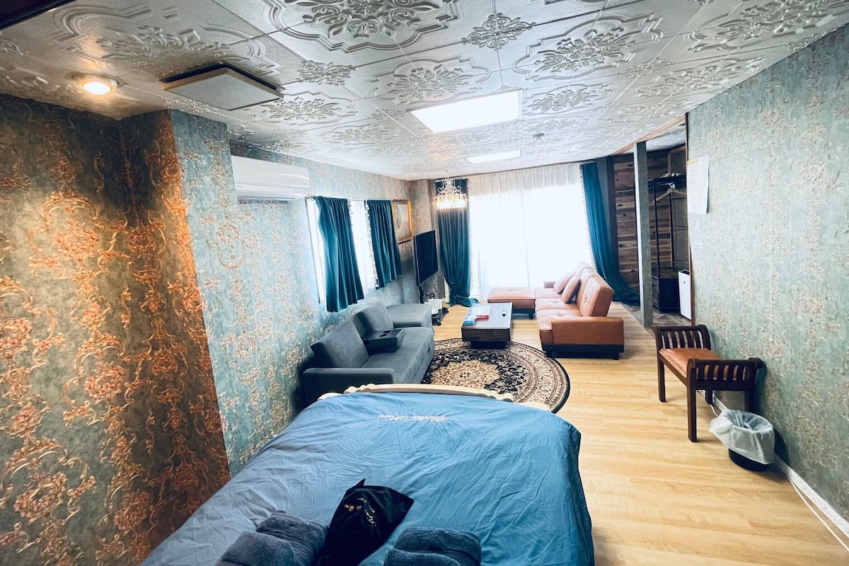 A room with a bed, a sofa, a small table, and a chair. There is a window with curtains and a chandelier.