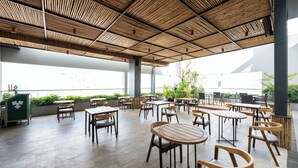 Un café en plein air avec des tables et des chaises en bois, un plafond en bambou et de grandes fenêtres donnant sur un espace verdoyant.