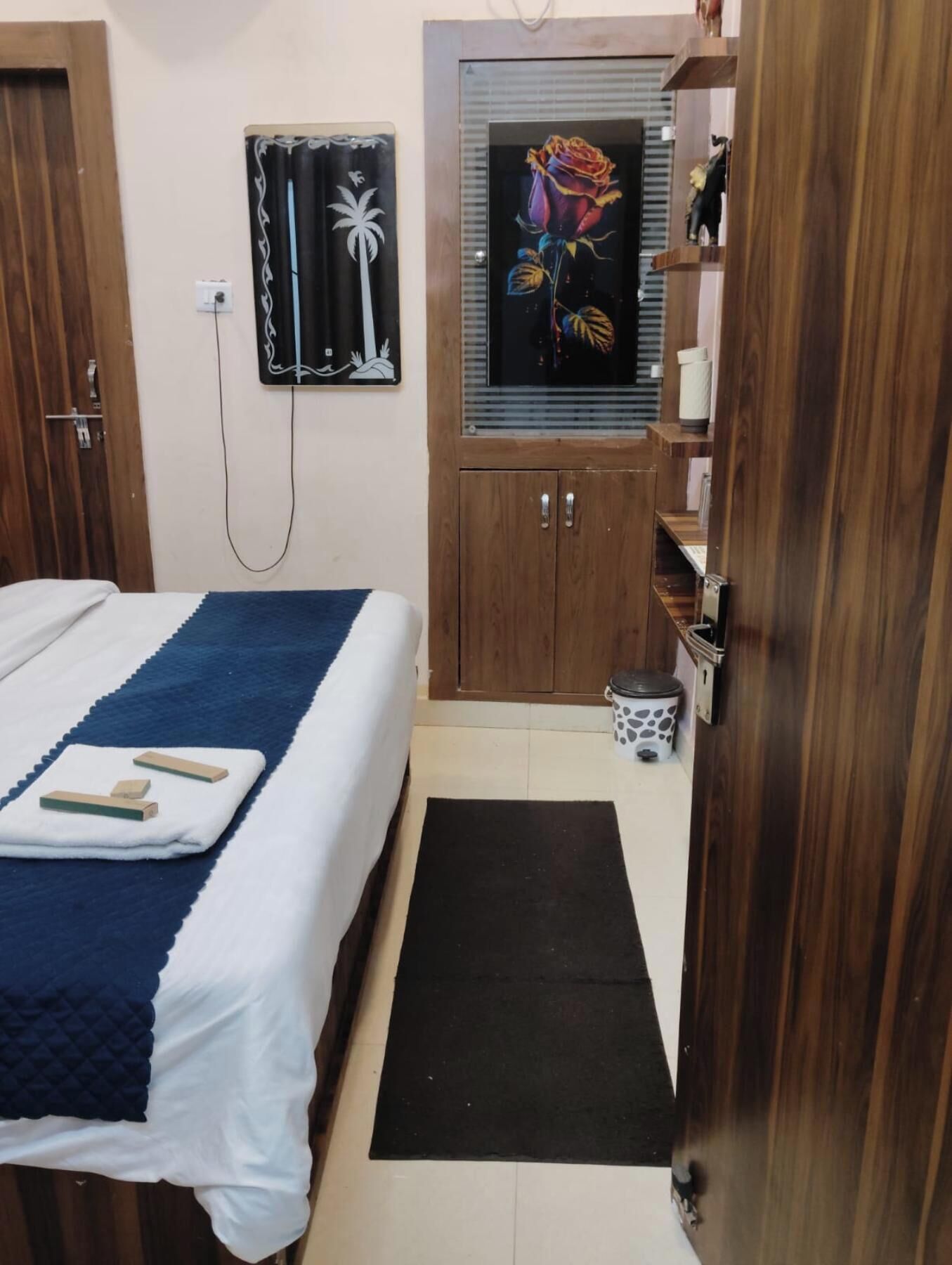 A bedroom with a bed, a wooden wardrobe, a small cabinet, a painting, and a door.