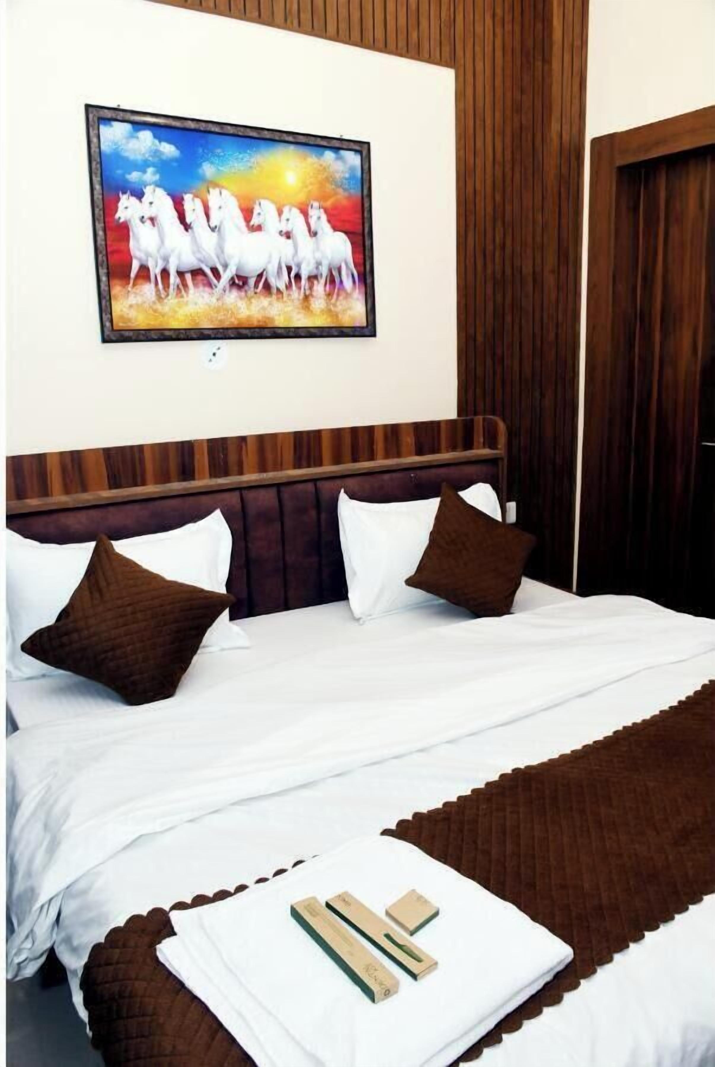 Una habitación de hotel con cabecera de madera, ropa de cama blanca, almohadas marrones y una manta marrón. En la pared, un cuadro de caballos blancos.