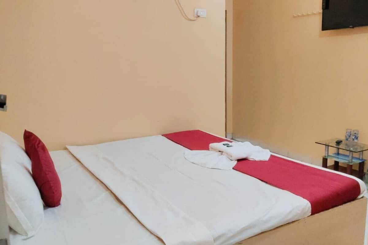 A single bed with a white and red bedspread, a red pillow, and a folded white shirt on the bed. There is a small table with a glass top and a few items on it, and a television mounted on the wall.
