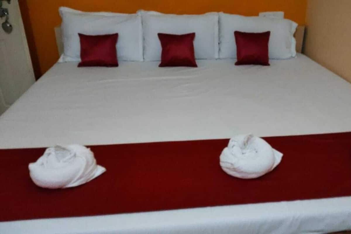 A hotel room with a bed, white bedding, red pillows, and three towels placed on a red bed runner.