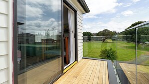 Een balkon met een glazen deur naar een kamer, een houten terras en uitzicht op een groen gazon en bomen.