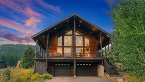 A large wooden cabin with a balcony, a garage, and a staircase leading to the upper level. - Alaskan Moose Lodge | Yellowstone + Hot Tub, Fire Pit & AC (Island Park)