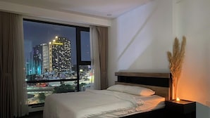 Un dormitorio moderno con una cama grande, una mesita de noche con una vela y vistas a un paisaje urbano a través de un ventanal.