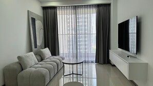 Una sala de estar moderna con un sofá gris, una mesita redonda pequeña, un televisor de pantalla plana montado en la pared y cortinas de gasa sobre un ventanal grande.