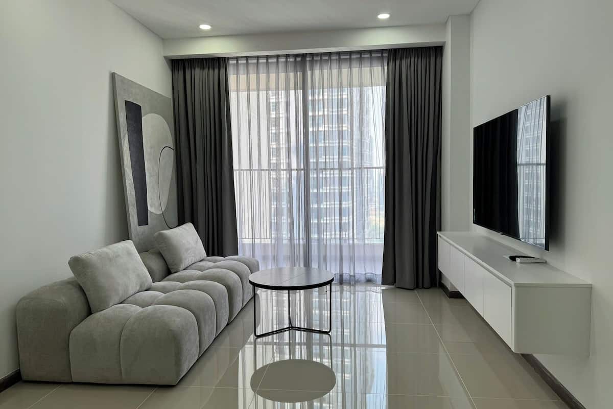 A modern living room with a grey sofa, a small round table, a flat-screen TV mounted on the wall, and sheer curtains over a large window.