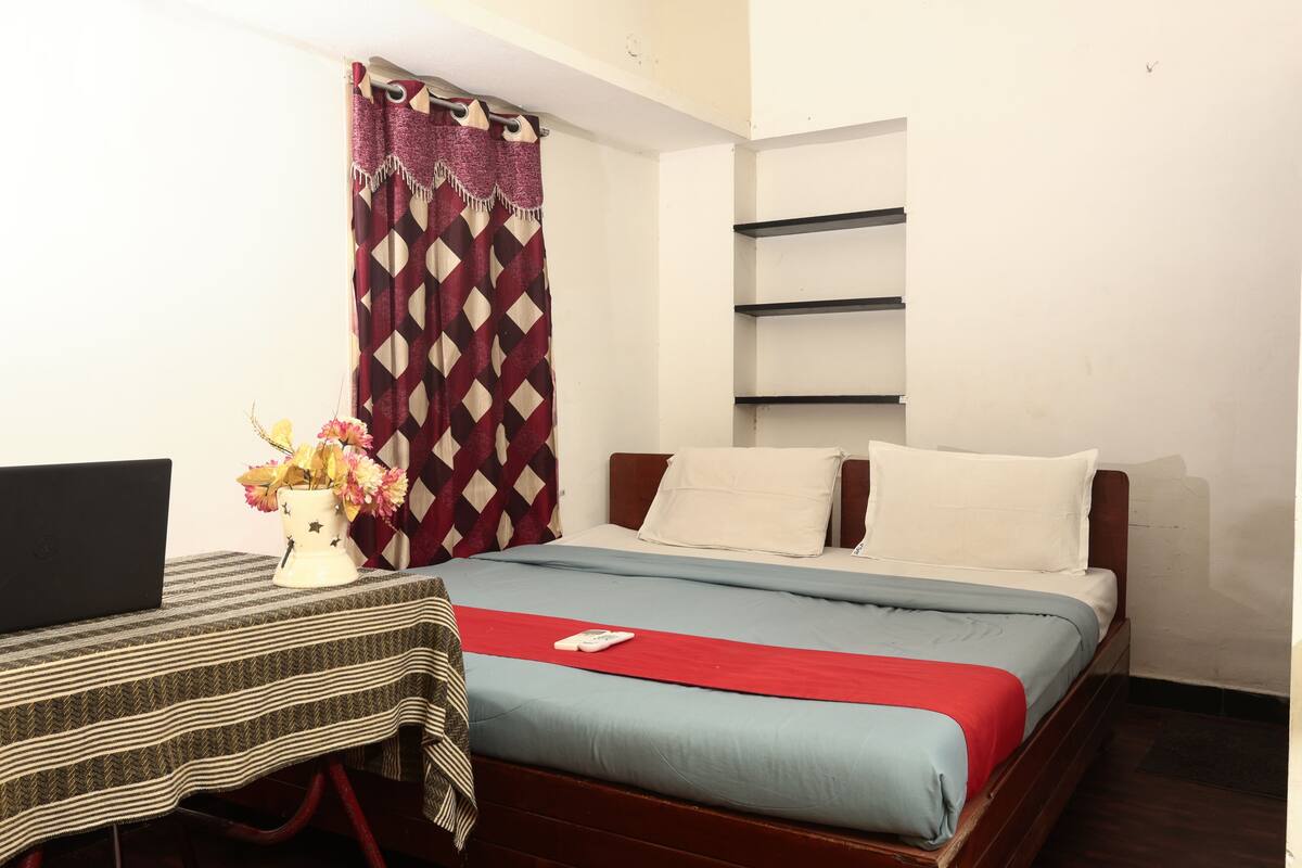 Habitación de hotel ordenada con una cama, un escritorio con una computadora portátil y un florero con flores.