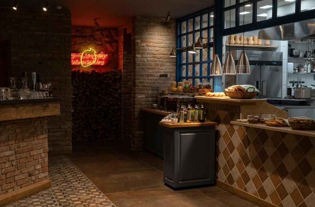 A restaurant interior with a brick wall, a neon sign, a kitchen area, and a counter with food items.