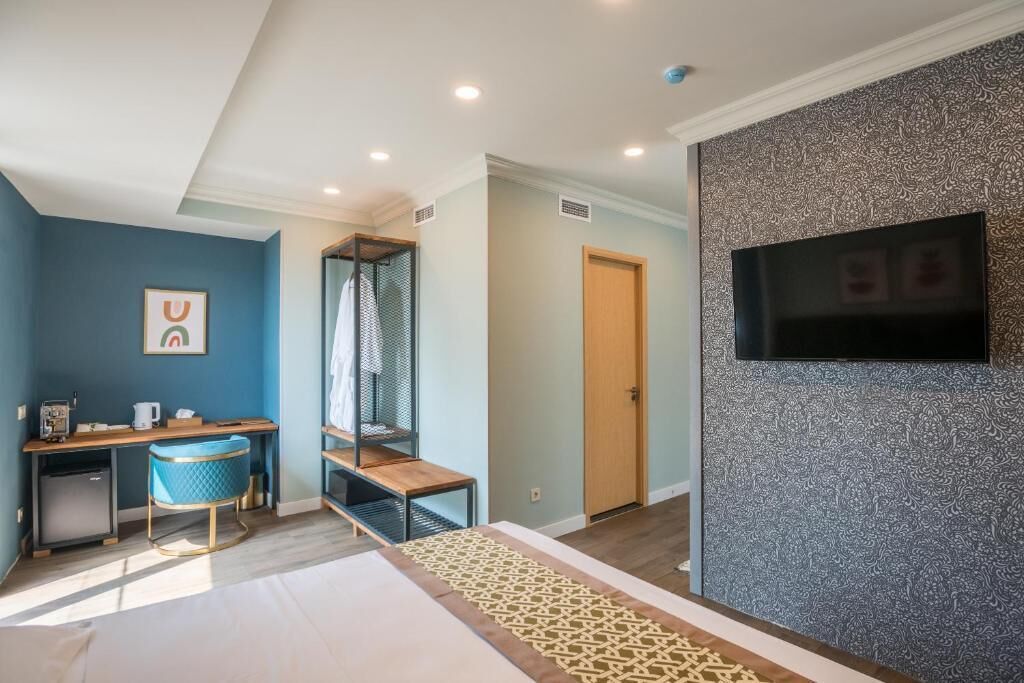 A modern hotel room with a bed, a desk, a chair, a TV, and a patterned wall.