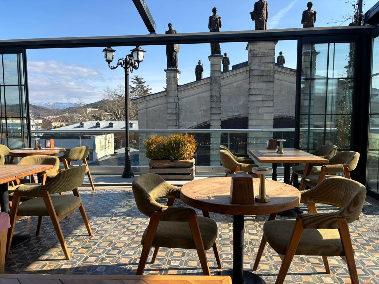 An outdoor seating area with tables and chairs, a potted plant, and a view of a building with statues.
