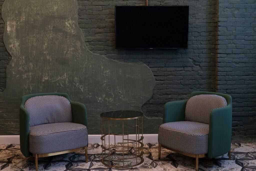 A room with two teal armchairs and a small round table, a flat-screen TV mounted on a brick wall, and a patterned carpet.