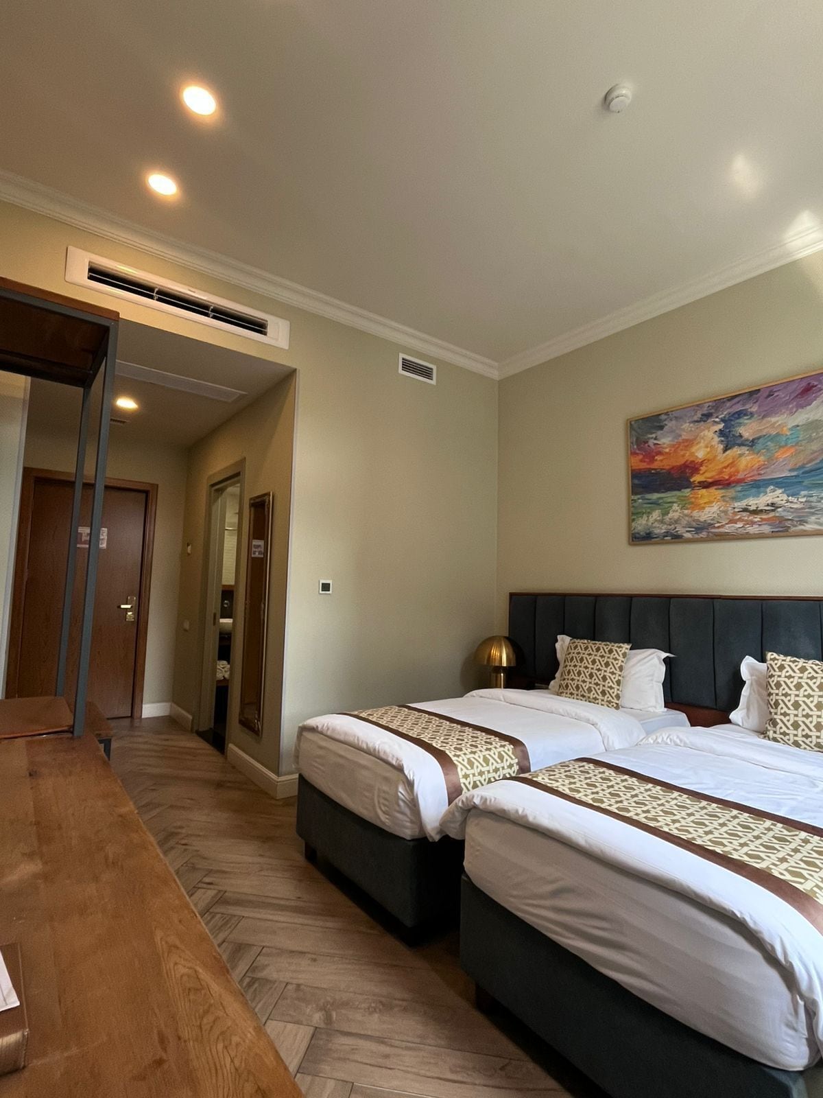 A hotel room with two beds, a wooden table, and a painting on the wall.