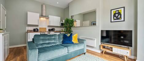 A modern living room with a teal sofa, a patterned rug, a flat-screen TV, and a kitchen area with white cabinets and a microwave.