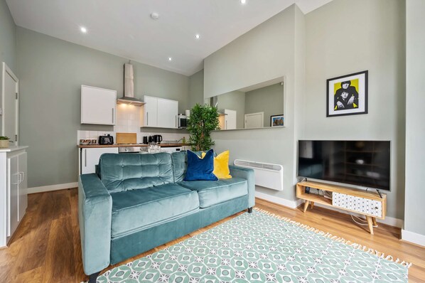 A modern living room with a teal sofa, a patterned rug, a flat-screen TV, and a kitchen area with white cabinets and a microwave.