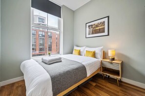 A modern hotel room with a large bed, a bedside table with a lamp, and a view of a cityscape through a large window.