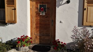 A wooden door with a wreath, a wall-mounted light, and two wicker baskets with red flowers. - Vacation apartment Rehkitz (Reit im Winkl)