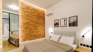 A modern bedroom with a brick accent wall, a large bed with a headboard, bedside tables, and a mirror. - G Residence - Luxe 1-bed, smart home on Ermou (Athina)