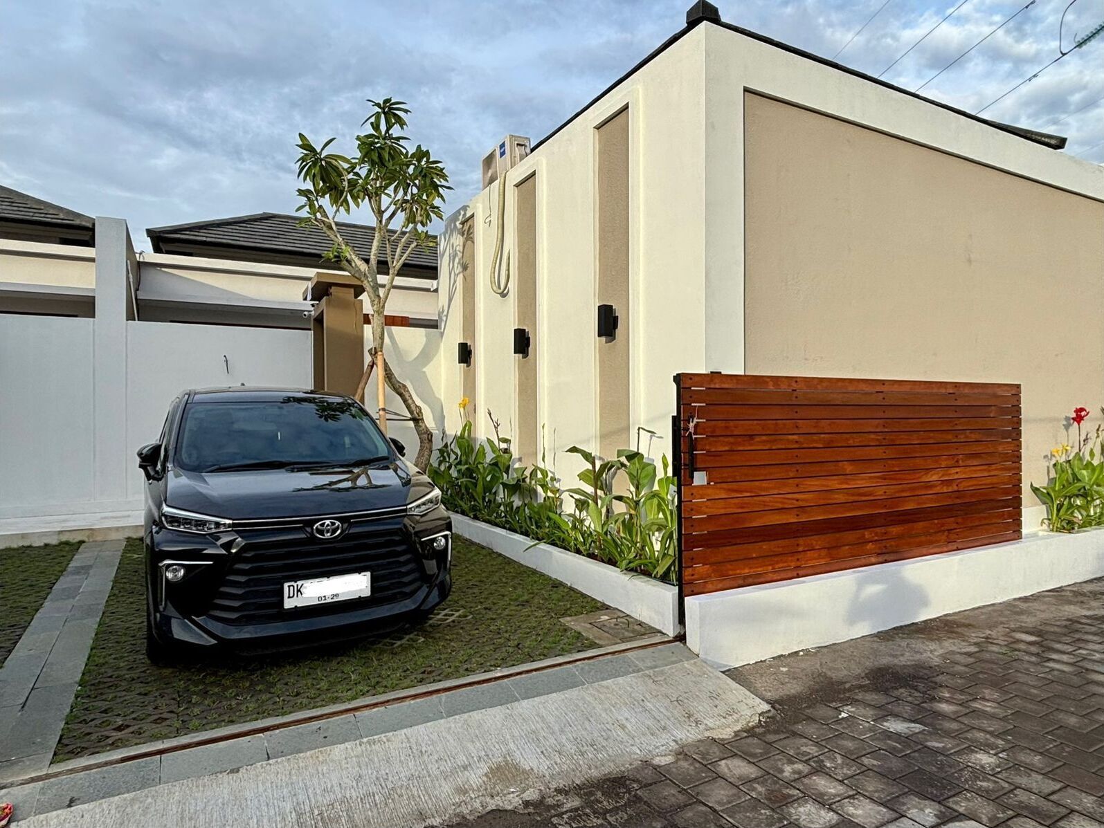 A modern house with a black car parked in the driveway, a wooden gate, and a small garden with plants.