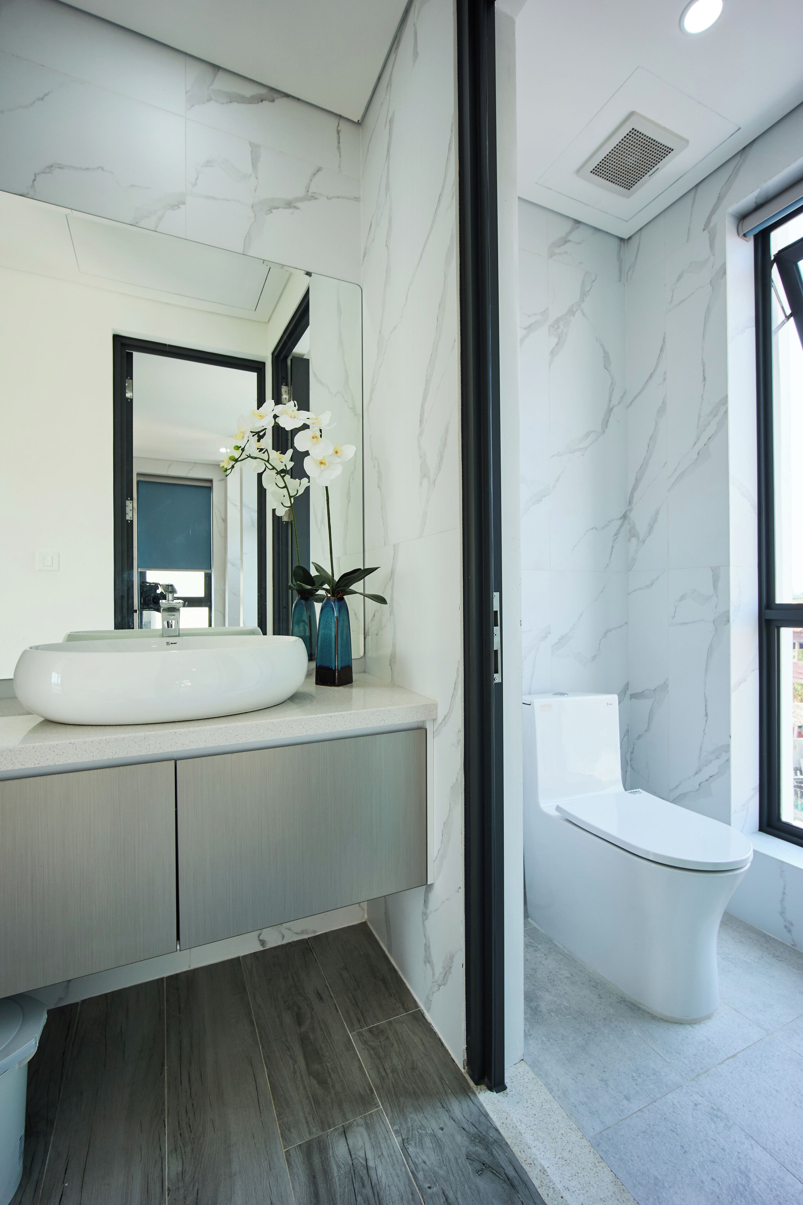 A modern bathroom with a white marble wall, a round sink, a blue vase with flowers, and a white toilet.