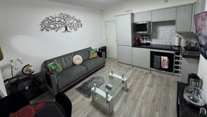 A modern living room with a grey sofa, a glass coffee table, and a kitchen area with white cabinets and a microwave. - Cozy 2br-5guests-near Attractions-garden (London)