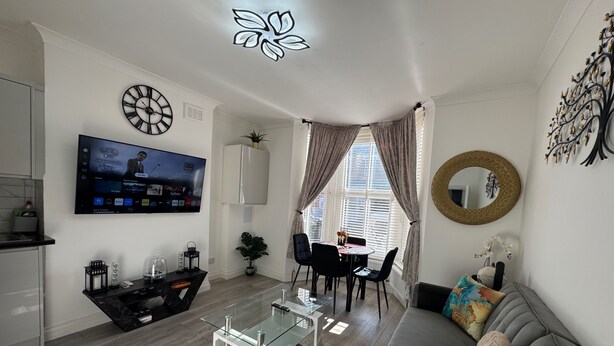 A modern living room with a flat-screen TV, a glass coffee table, a gray sofa, and a dining area with a table and chairs. - Cozy 2br-5guests-near Attractions-garden (London)