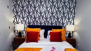 A hotel room with a geometric patterned wall, a bed with orange and white bedding, two nightstands with lamps, and a decorative pillow with a flower.