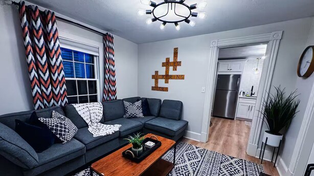 A modern living room with a sectional sofa, a wooden coffee table, patterned rugs, and a kitchen in the background.
