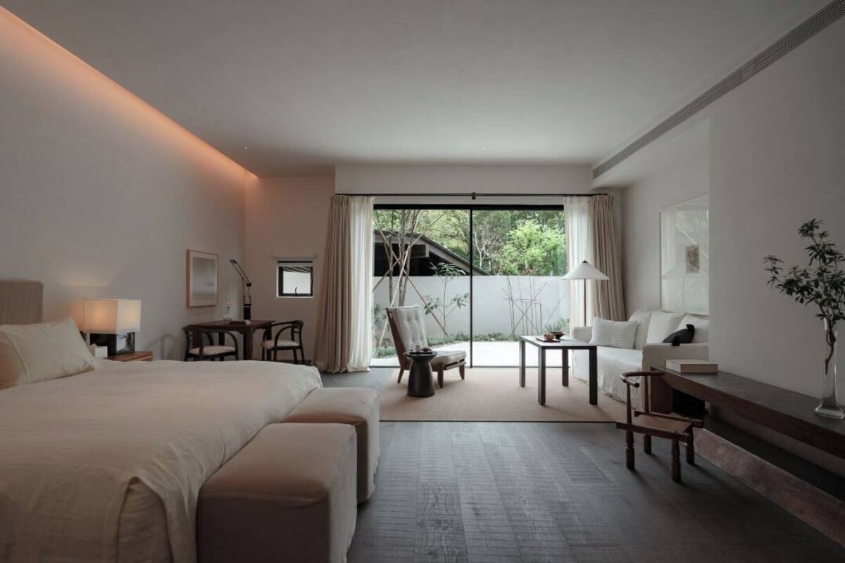 A hotel room with a large bed, a desk, a chair, and a view of the outdoors through a large window.