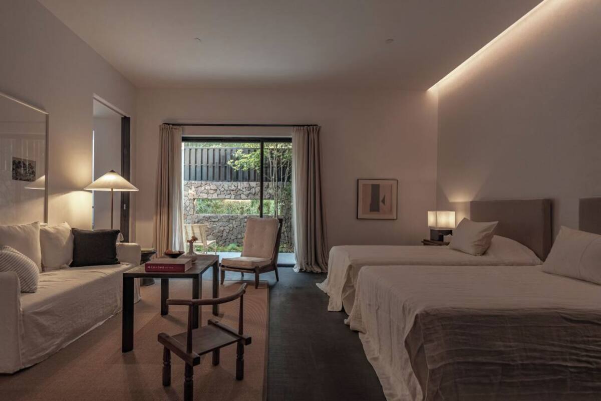 A hotel room with two beds, a sofa, a table, and a chair. There is a window with curtains and a view of greenery outside.