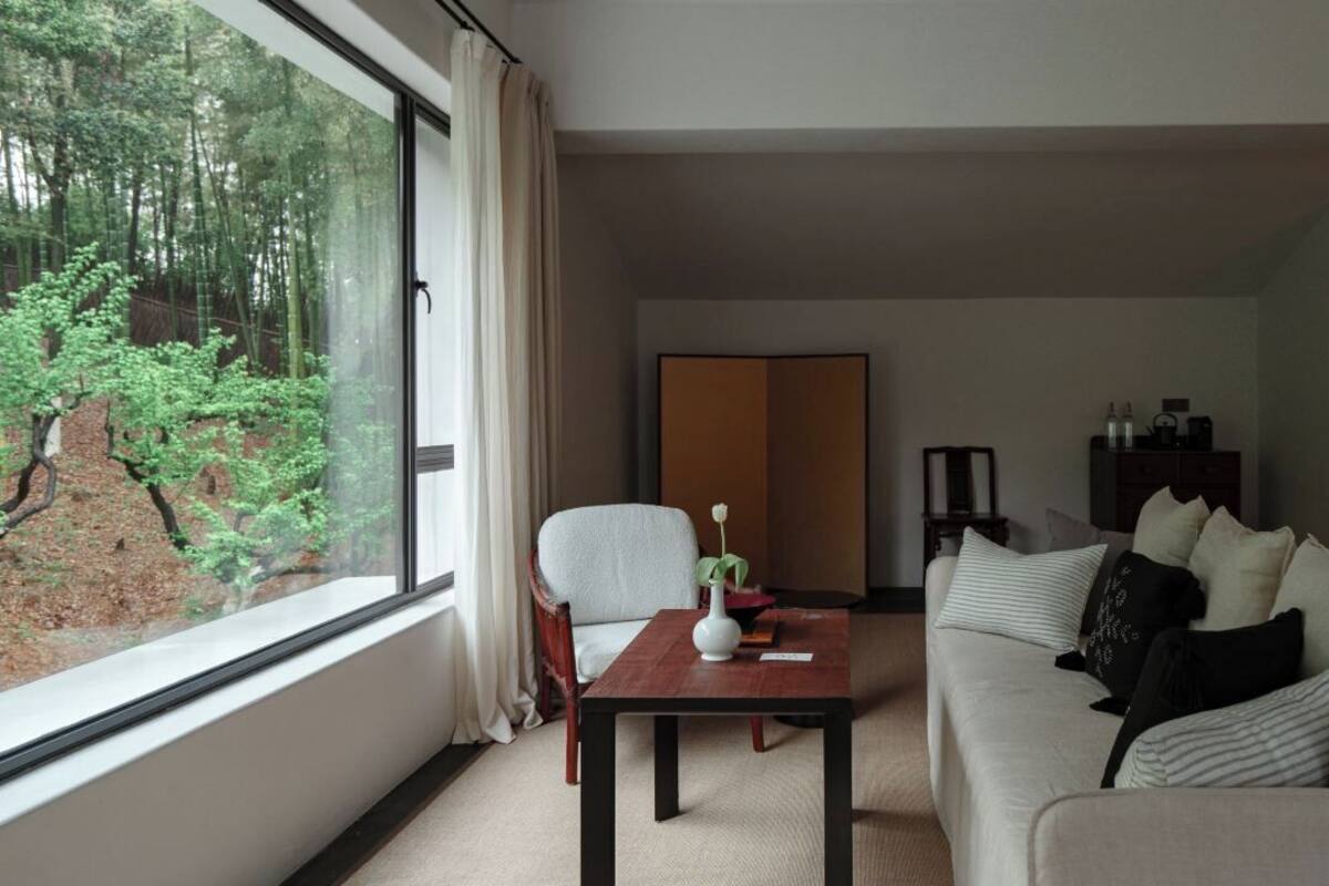 A modern hotel room with a large window offering a forest view, a sofa, a wooden coffee table with a vase, and a traditional Japanese sliding door.