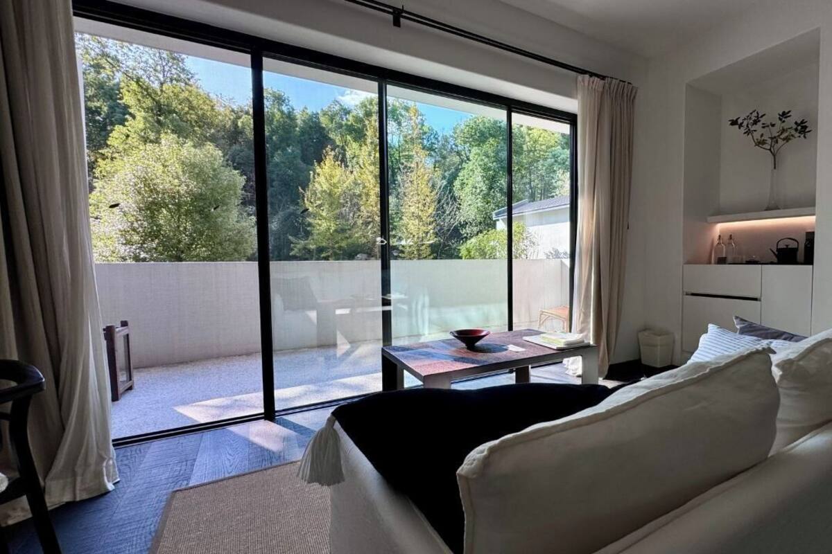 A modern room with a large sliding glass door leading to a balcony, a white sofa, a small table with a bowl, and a shelf with decorative items.