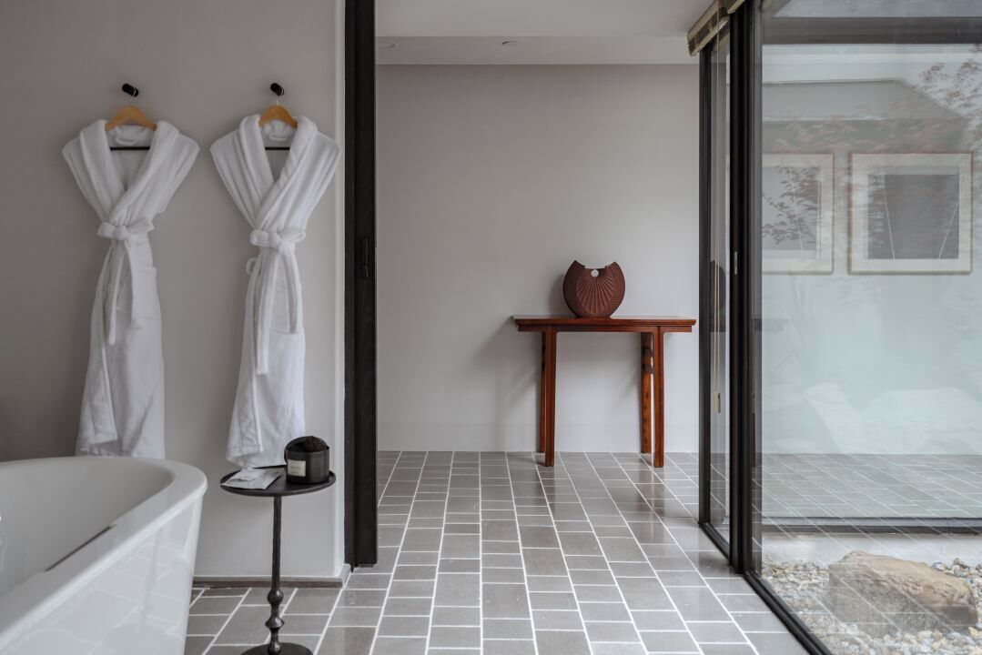 A modern bathroom with a bathtub, two white robes hanging on a rack, a small table with a vase, and a view through a glass door.