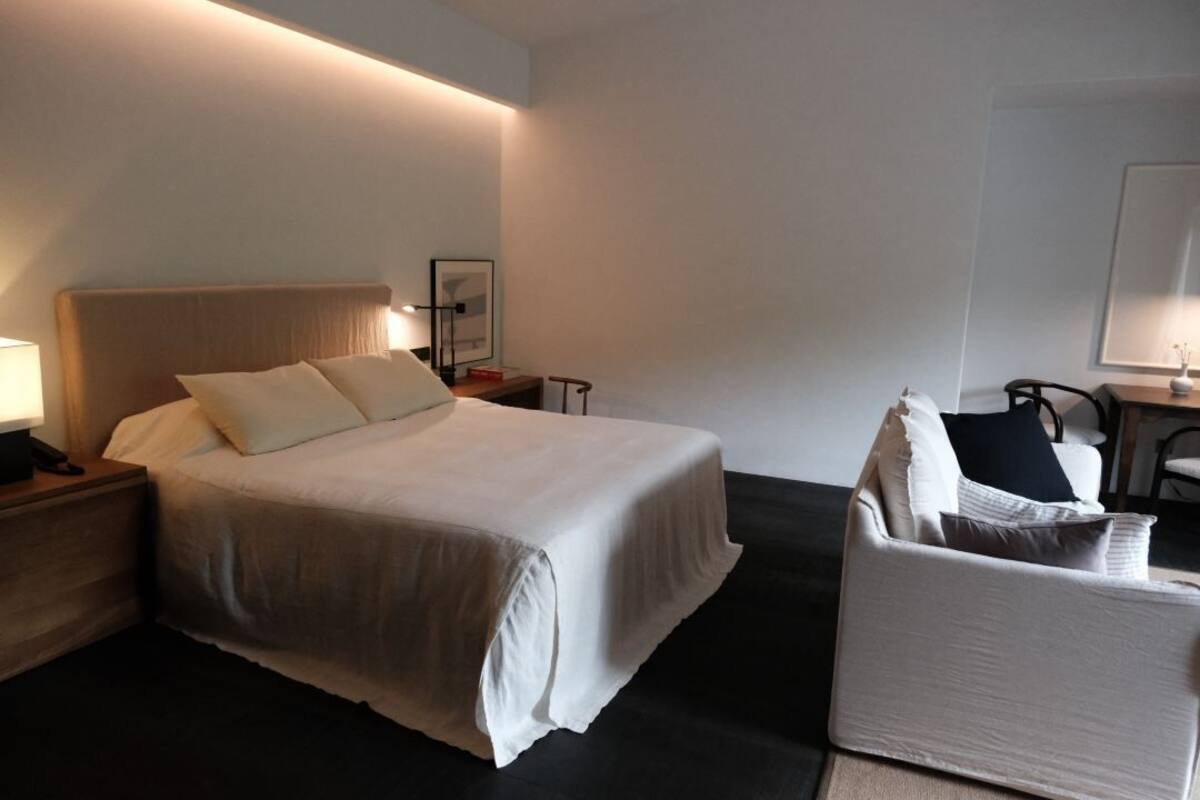 A hotel room with a large bed, a bedside table with a lamp, a chair, and a small table with a vase.