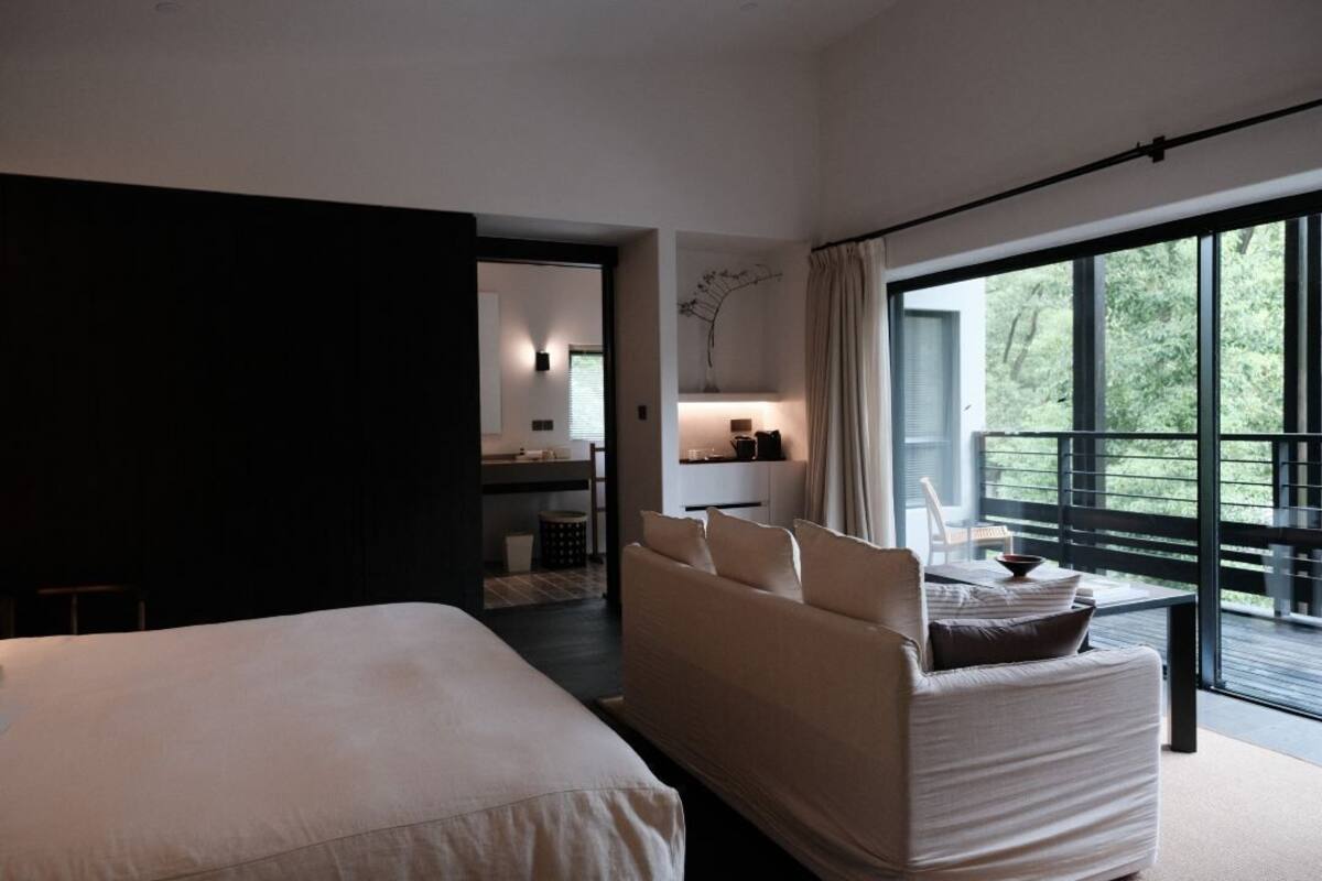 A modern hotel room with a large bed, a sofa, a balcony with a view, and a small kitchenette.