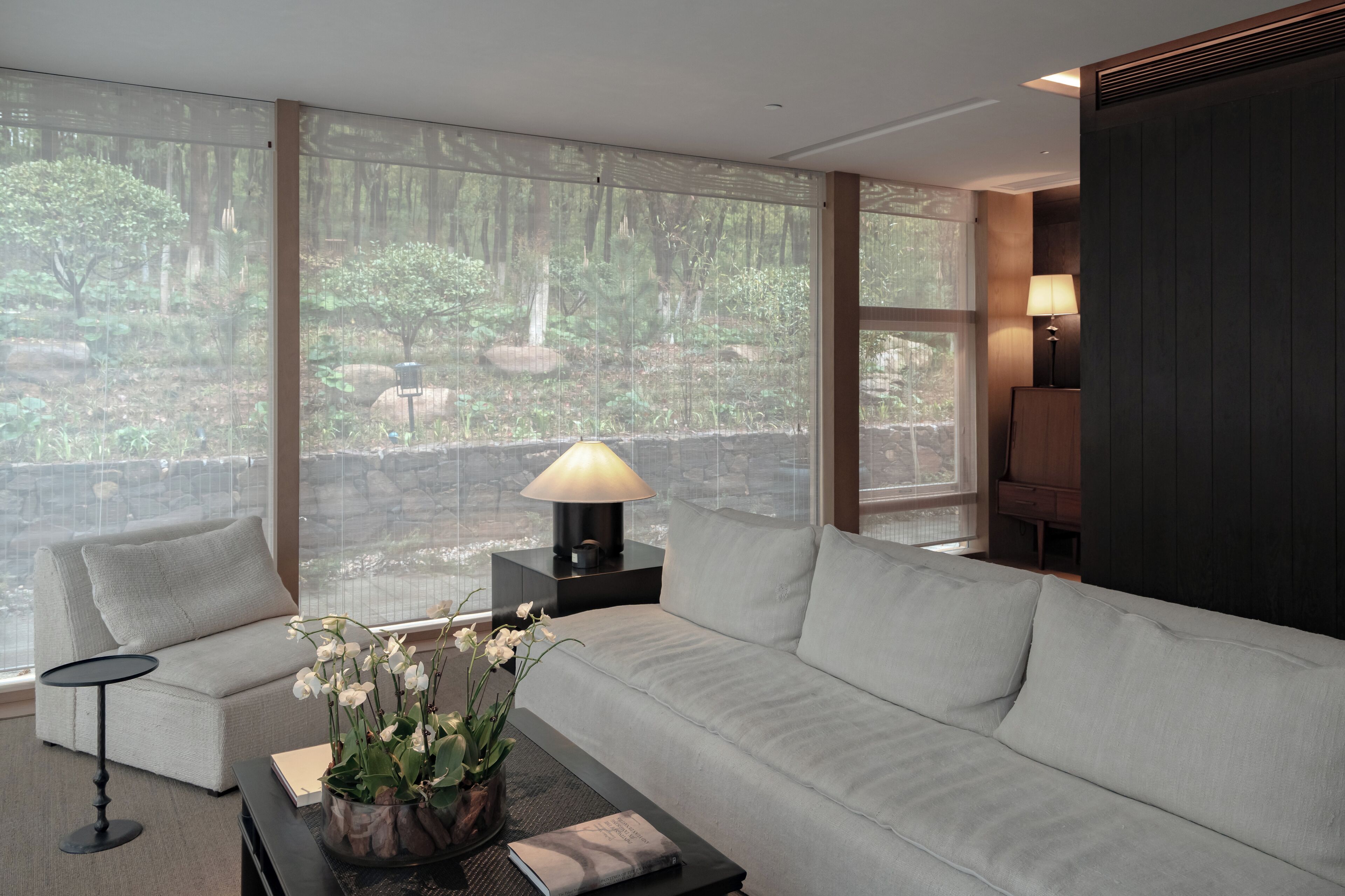 A modern living room with a large window, a sofa, a coffee table with a lamp, and a view of a forest.