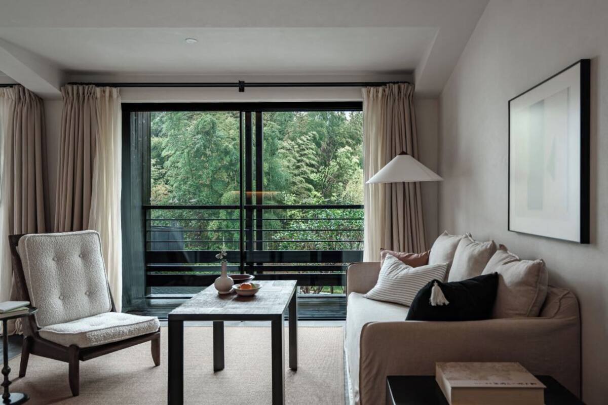 A modern living room with a sofa, armchair, and a table with a vase and book. A large window offers a view of greenery outside.