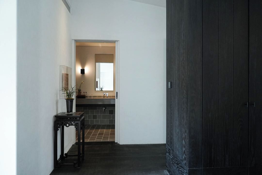 A hallway leading to a kitchen with dark wooden flooring, a black table with a plant, and a white wall with a light fixture.