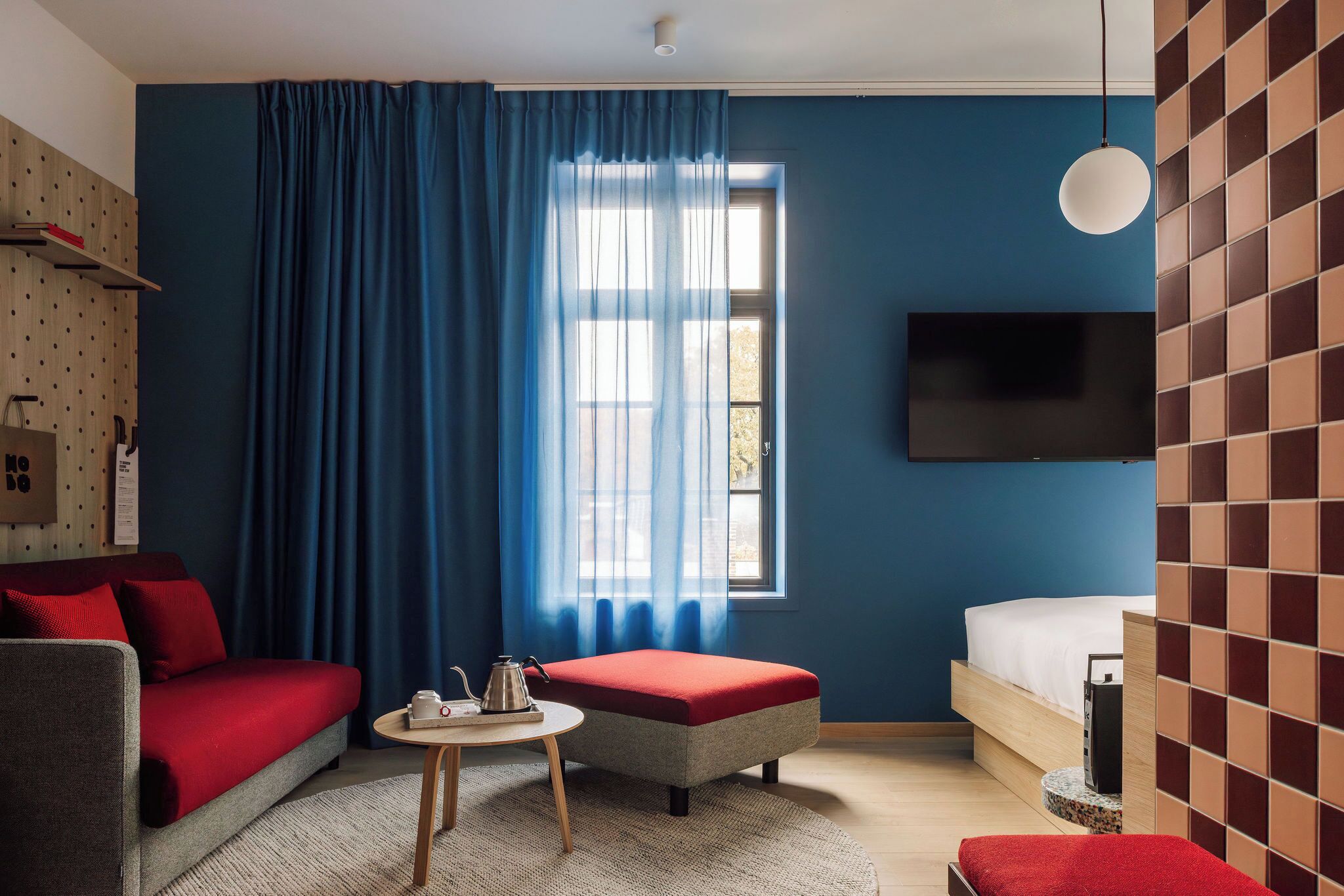 A modern hotel room with a blue wall, a large window with blue curtains, a grey sofa with red cushions, a small round table with a kettle, a red ottoman, a bed with white bedding, and a mounted TV.