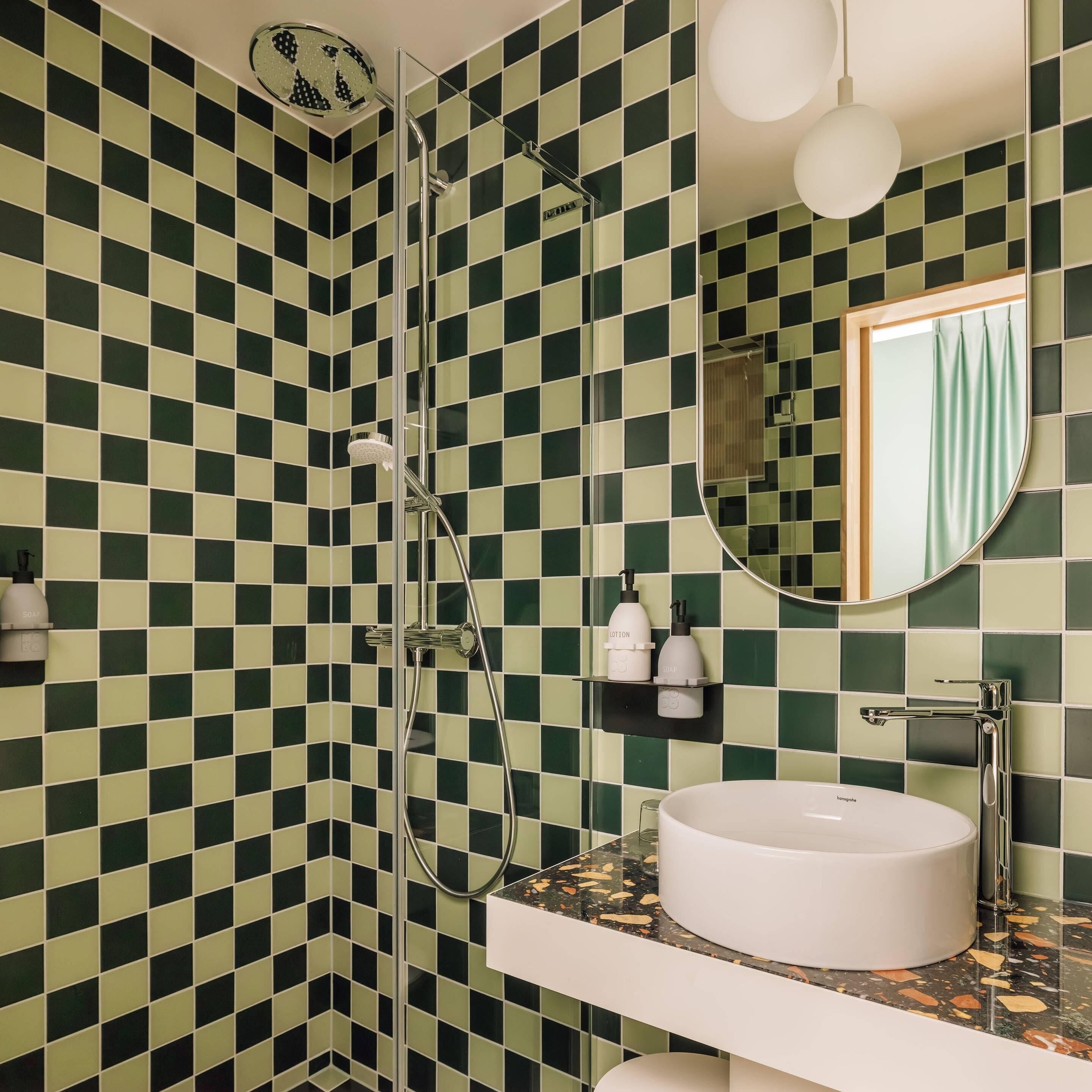 Um banheiro com parede de azulejos quadriculada em verde e branco, chuveiro com porta de vidro, pia branca com bancada de mármore e espelho redondo com cortina verde.