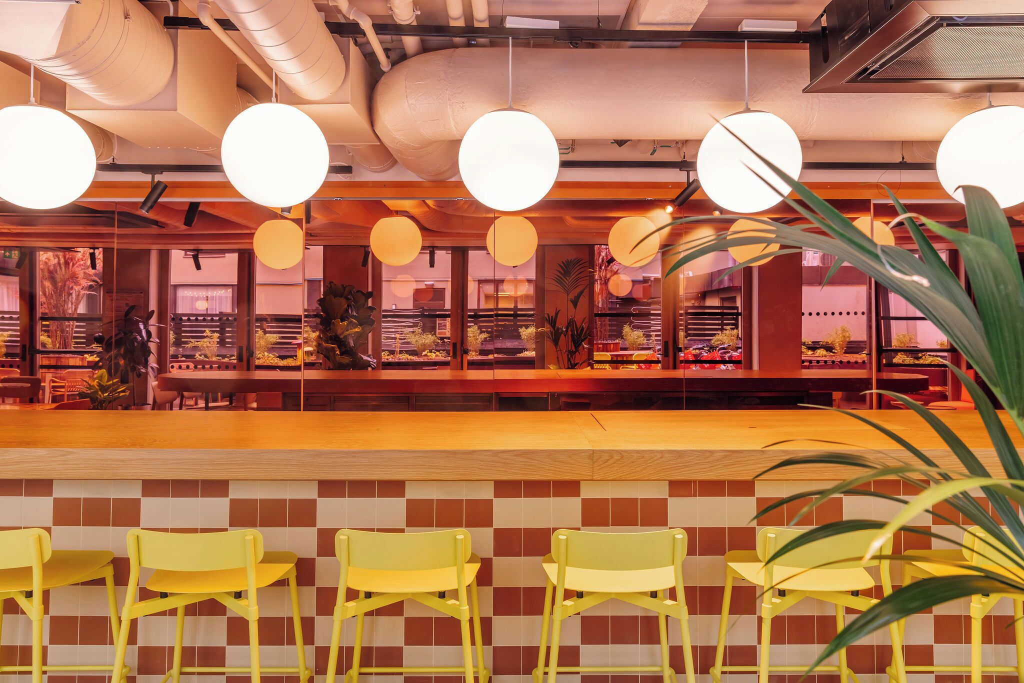 Interior de restaurante com parede de azulejos quadriculados, cadeiras amarelas e luminárias esféricas penduradas.