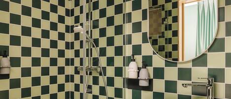 Un baño con pared de azulejos blancos y verdes a cuadros, una ducha con cabezal de mano, un lavabo redondo sobre encimera de mármol y un espejo con marco circular.