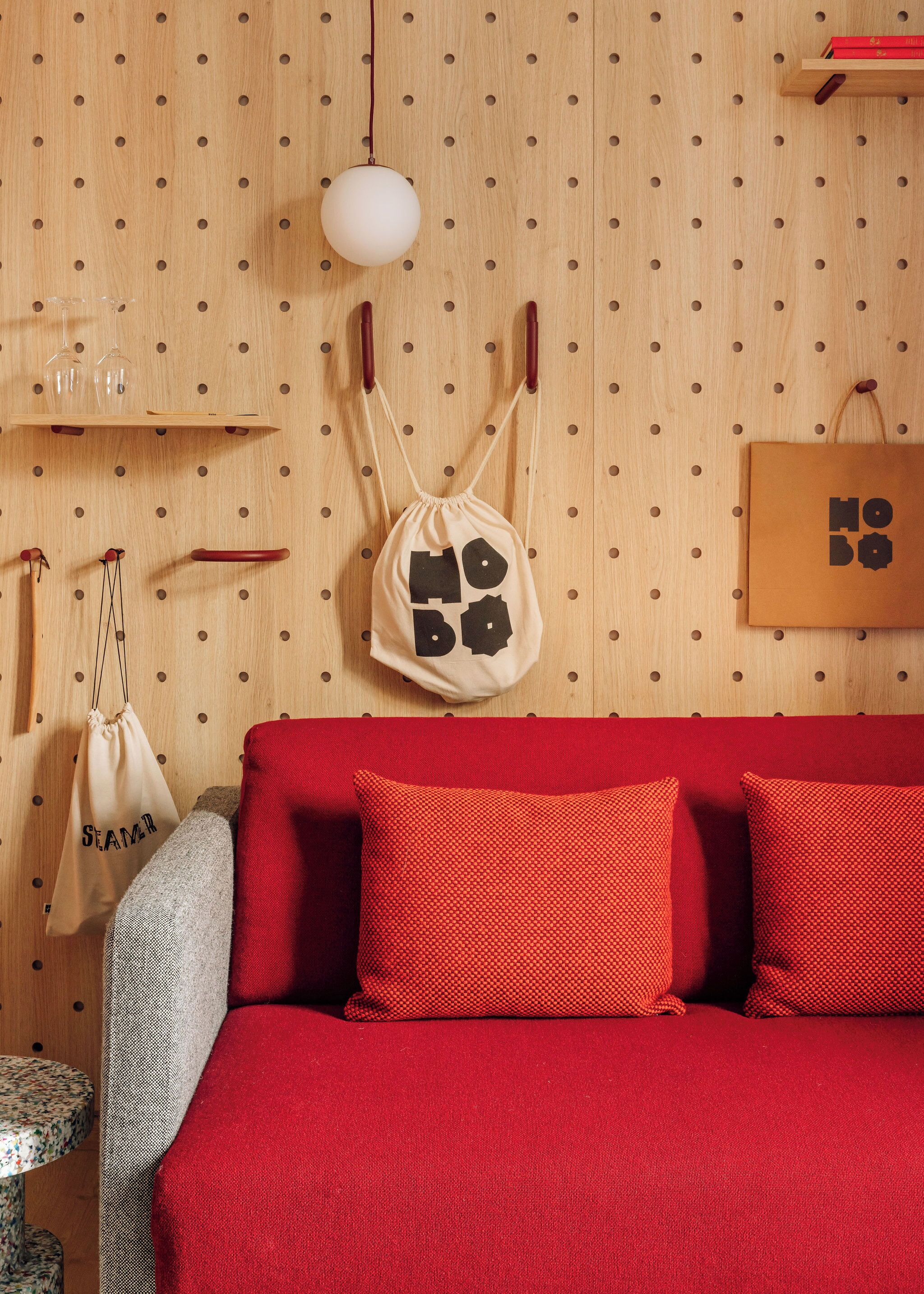 Ein Zimmer mit einem roten Sofa, einem gemusterten Sessel, einer Holzwand mit einem Nagelbrett und einer Pendelleuchte.