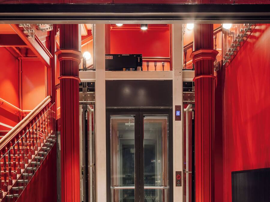 A red-walled elevator entrance with a glass door, a staircase to the left, and a red pillar on the right.