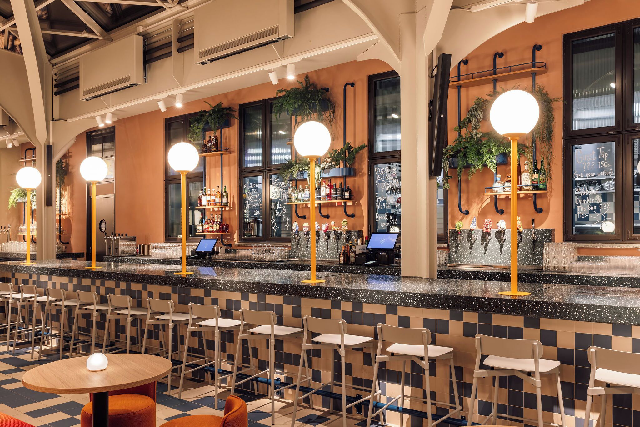 Uma área de bar com piso xadrez, banquetas e mesas redondas.
